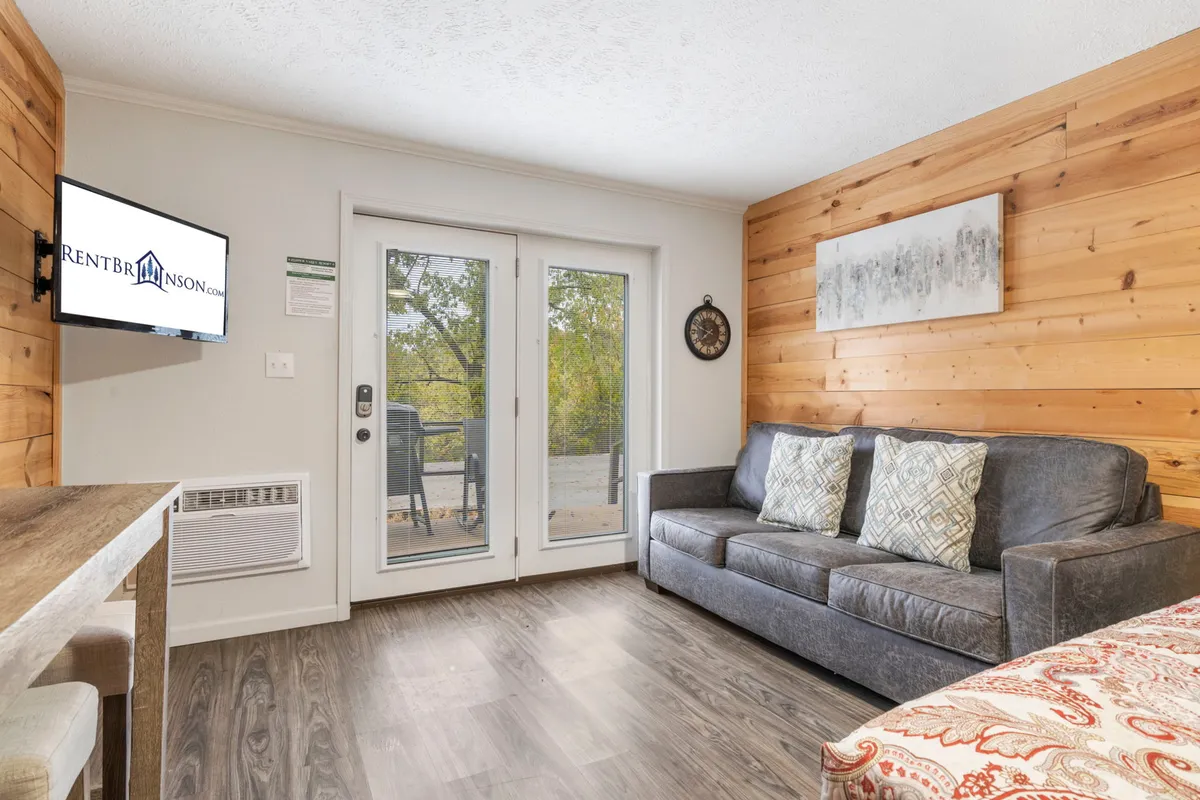 A warm, intimate living area with a comfortable sofa facing a television, complemented by sliding doors and a wooden accent wall. The natural morning light streams in, and the layout invites guests to relax, watch TV, or enjoy views outside.