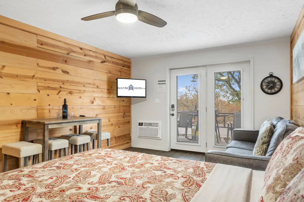 This image shows an inviting indoor space with a wooden accent wall, a cozy bed with patterned bedding, a small dining table, and a sliding glass door leading directly to a private patio. The view through the glass hints at the outdoor sanctuary, seamlessly connecting the interior to the patio and the lake beyond.