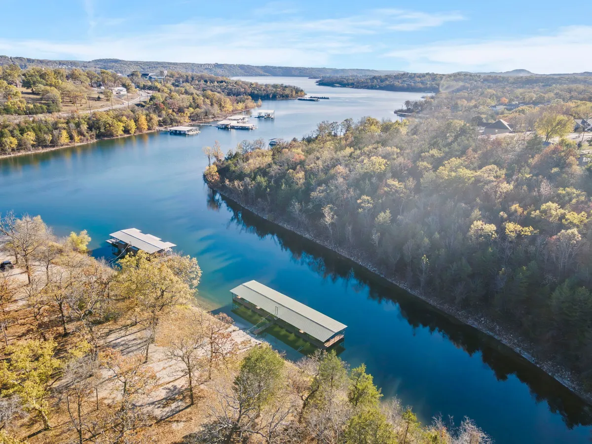This image captures a tranquil view across calm waters, featuring boathouses along a tree-lined shoreline and distant hills. People are engaged in boating and fishing, with the setting sun casting warm reflections on the lake.