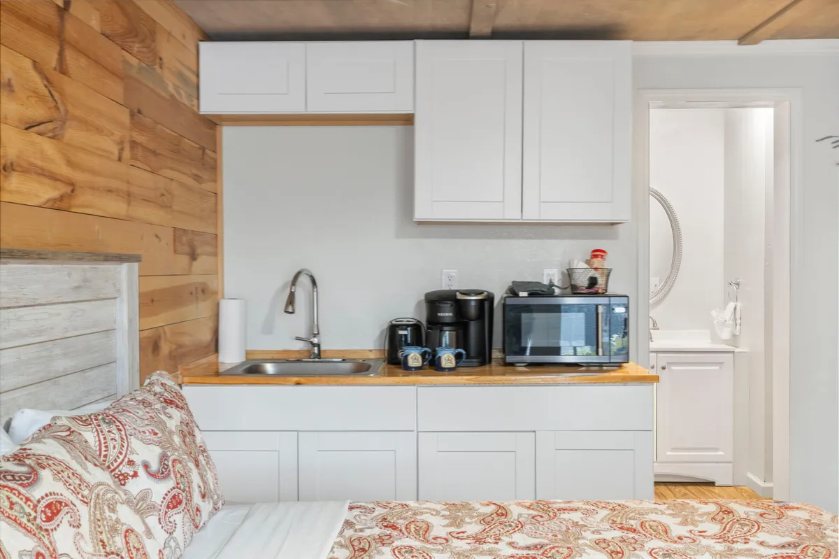 This interior scene combines a cozy bed adorned with decorative pillows, a clearly visible kitchenette area with a microwave and mini-fridge, and a bathroom door, all within a compact, inviting layout. The kitchenette is stocked and ready for simple meal prep, and the whole space exudes warmth and comfort.