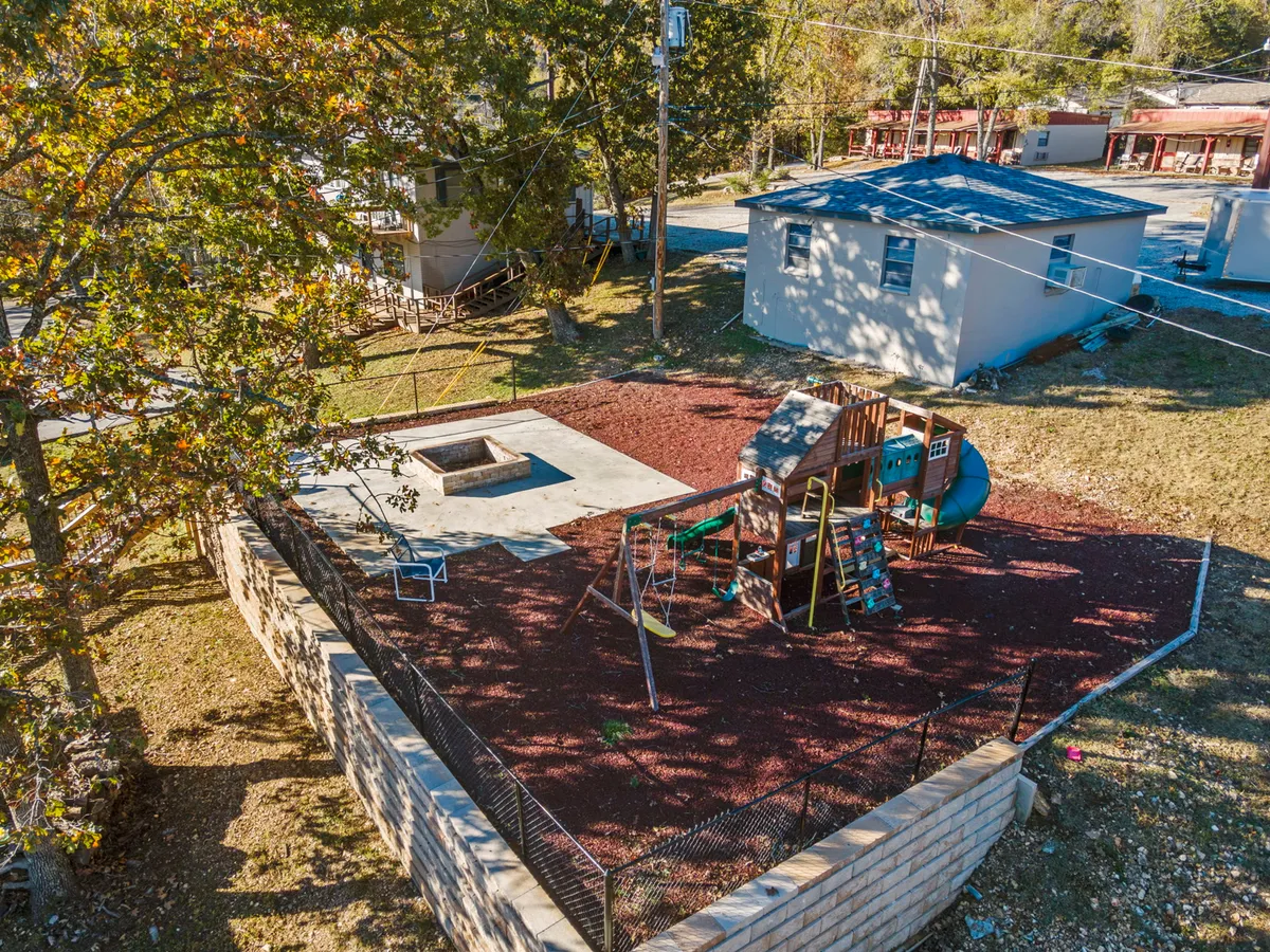 A playful outdoor playground scene with a colorful structure set in a sand pit, surrounded by trees and enclosed by a fence. The area is designed for children and family gatherings, evoking a sense of safety and joyful activity.