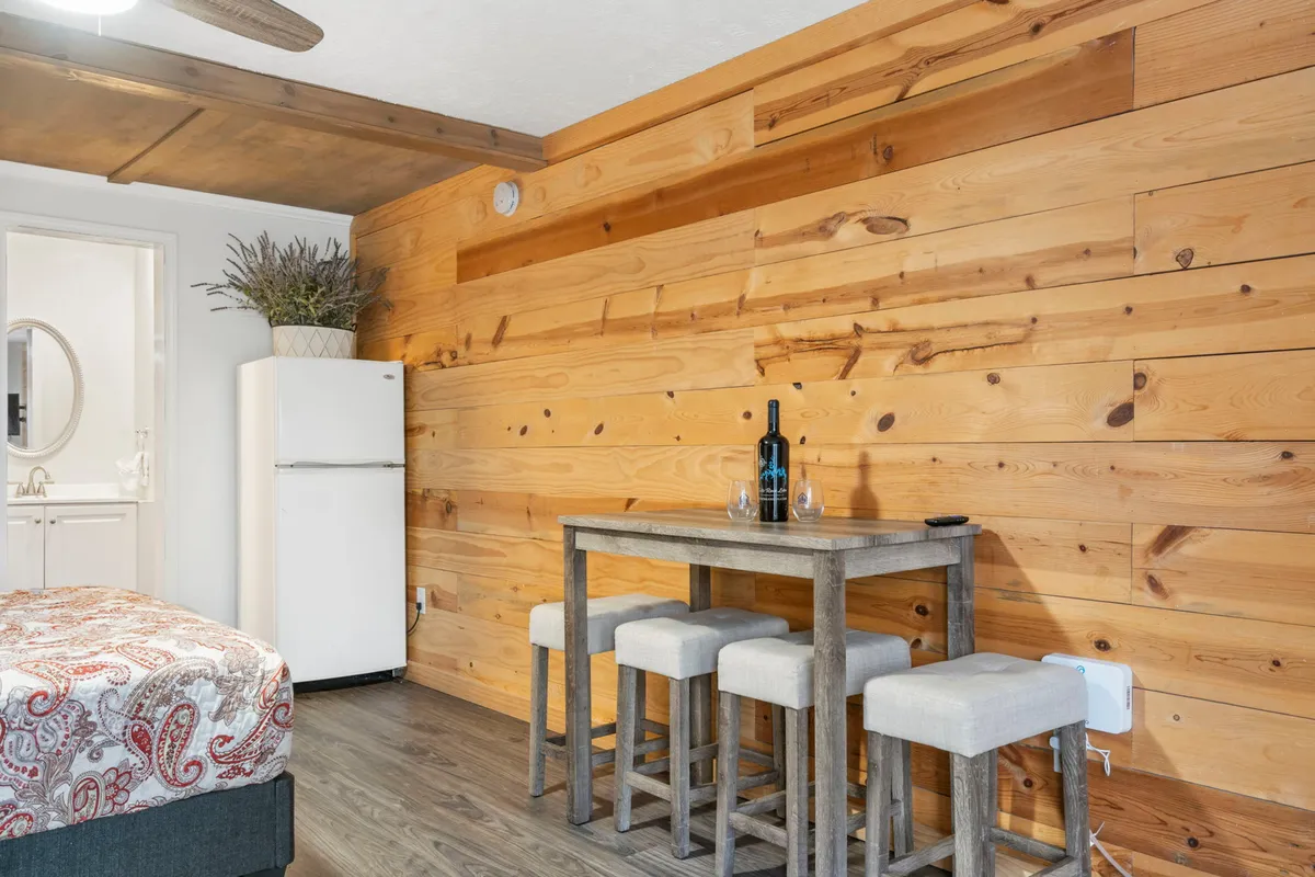 A cozy interior shot showcasing a small dining table with chairs, refrigerator, decorative plant, and bottle of wine against a wooden accent wall. The scene feels inviting, perfect for casual meals or unwinding after a day outdoors.