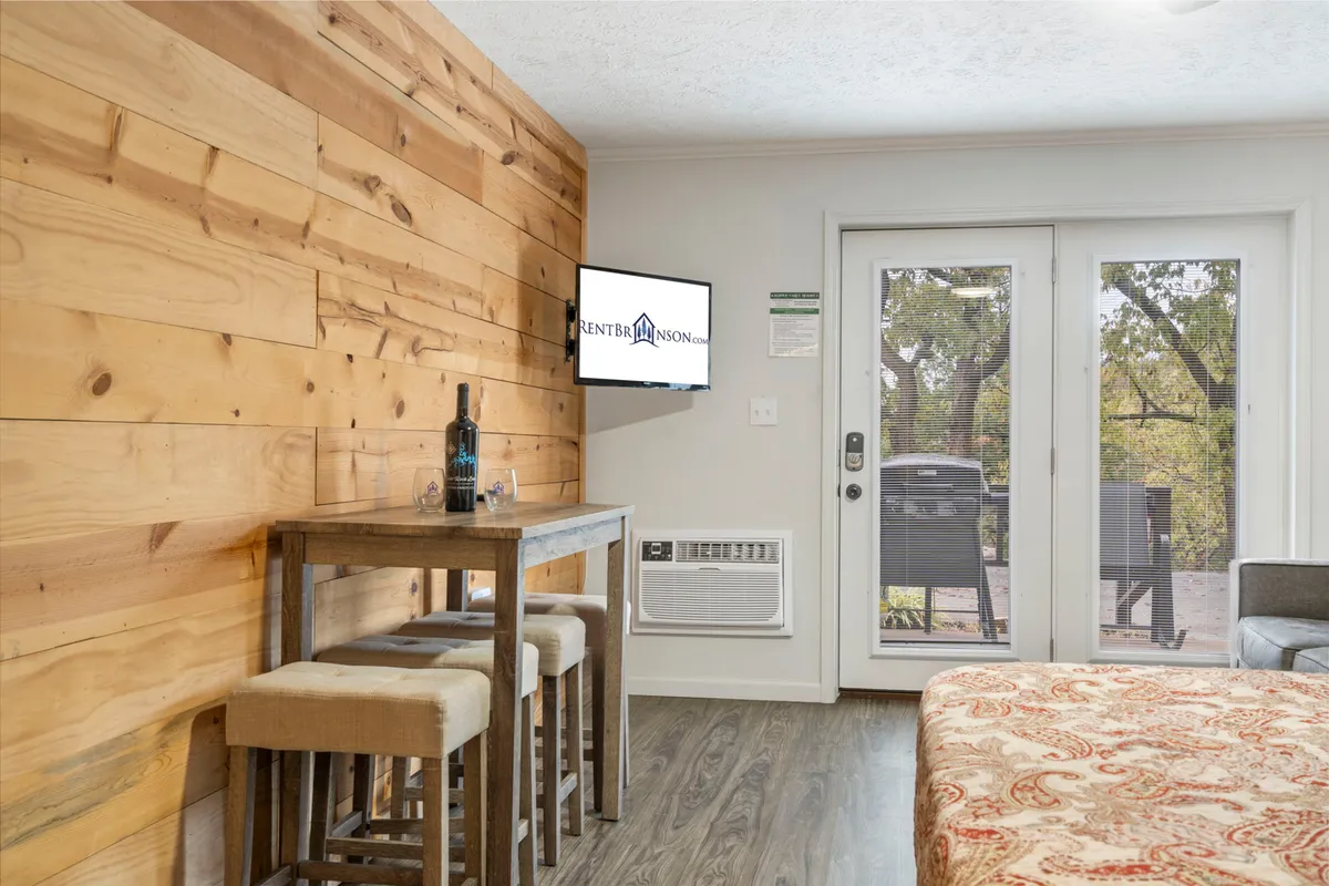 This image features a welcoming interior with a wooden accent wall, a bar table with stools, a television, and double doors that open out onto the patio. The setup suggests casual dining and relaxation with an easy transition to the outdoor escape.