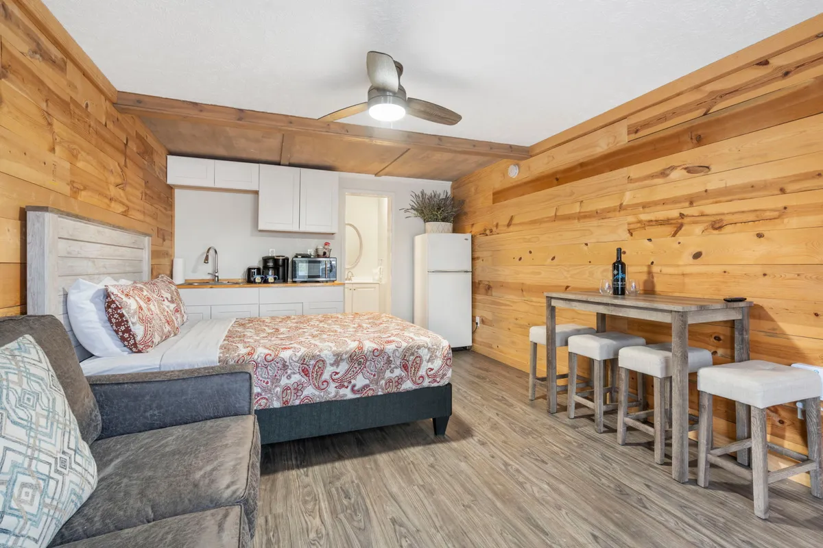 A warm, inviting interior with wood paneling, a small dining table with chairs, a patterned bed, and a visible refrigerator. The arrangement emphasizes the ease of sharing meals, relaxing, and socializing in a compact space.