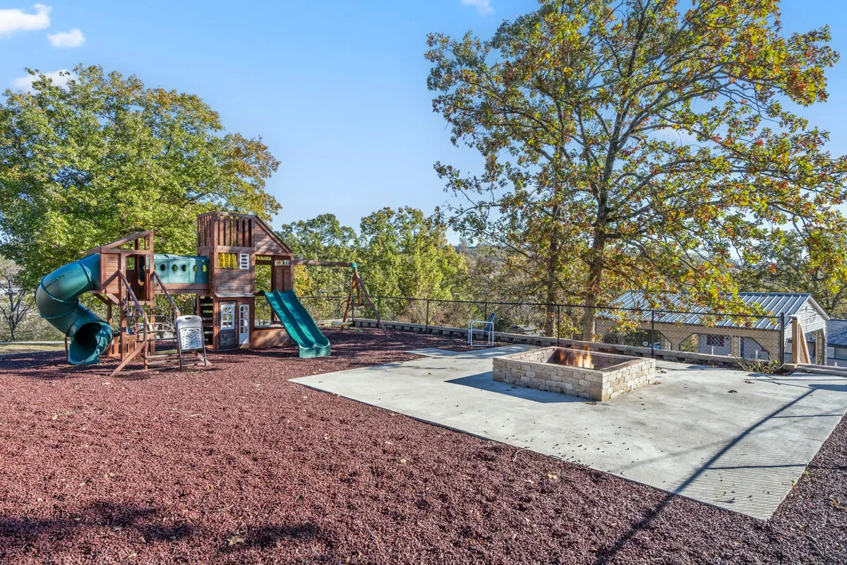 This exterior image features a vibrant playground structure adjacent to a communal firepit, surrounded by lush trees. The scene is lively, evoking the joy of children playing while families gather around the firepit, with the soft glow of sunset in the background creating a welcoming, communal mood.