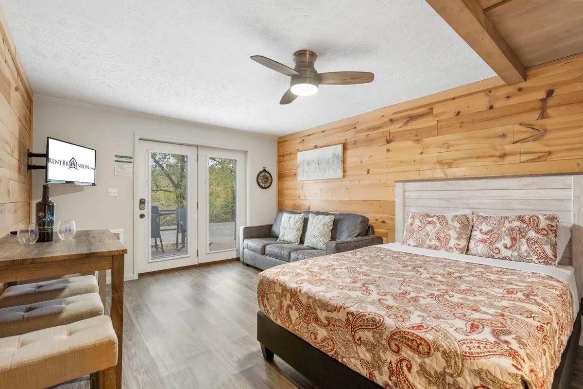 The image showcases a bright, inviting studio interior with a plush, patterned bed, a cozy sofa facing a wall-mounted TV, and large sliding glass doors that open to a balcony—hinting at lake views and abundant natural light.