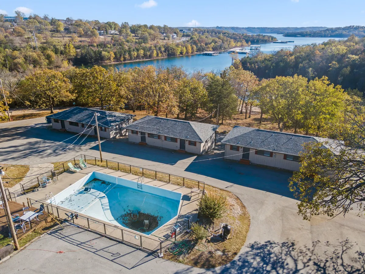 A serene exterior view featuring a swimming pool in the foreground, framed by lush trees and motel units, with a tranquil lake in the background. The scene hints at a variety of outdoor amenities and activities such as swimming, fishing, picnicking, and relaxing by the water.