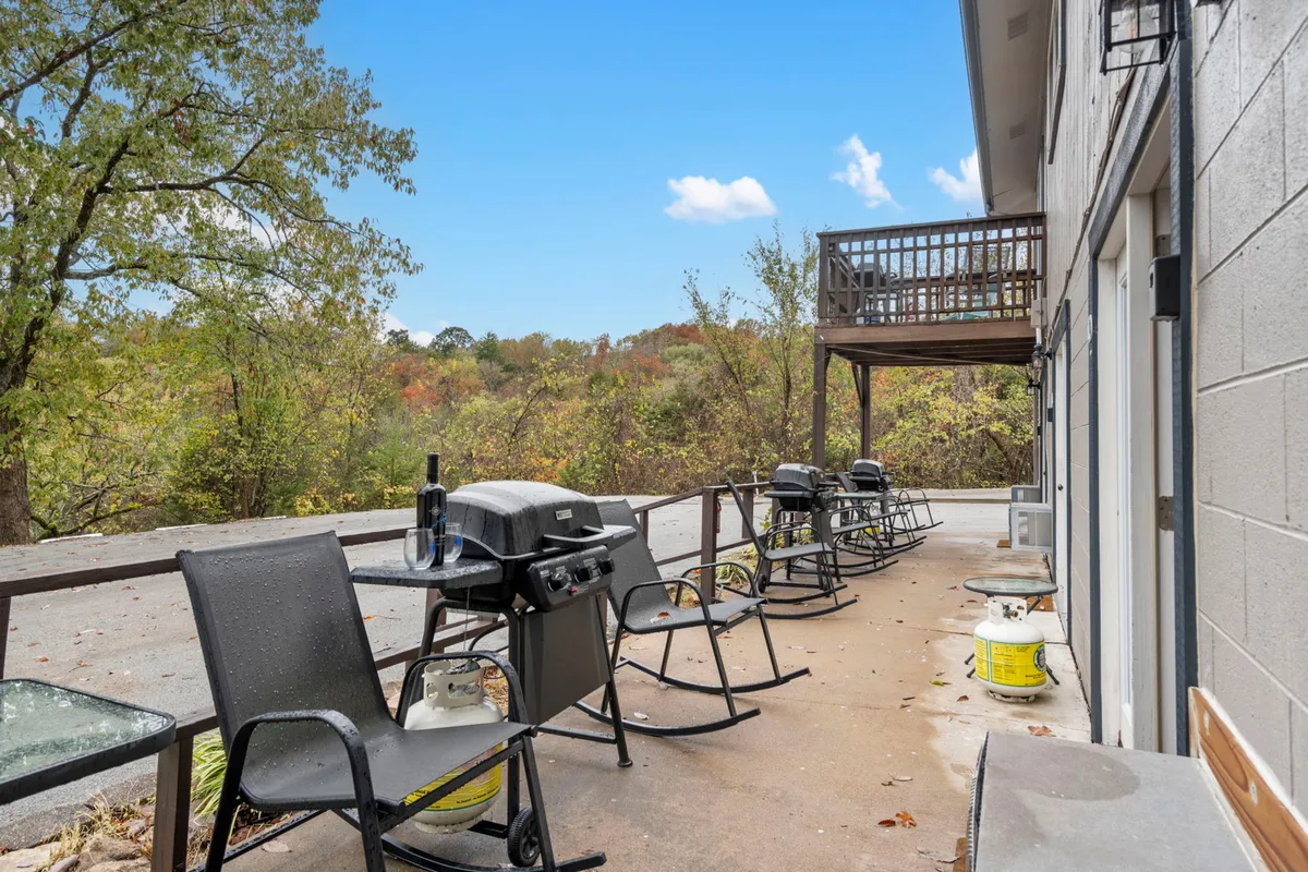 An exterior view showcasing a patio area with outdoor seating and a grill, surrounded by trees. The scene is relaxed, with hints of scenic views and a tranquil atmosphere—ideal for enjoying morning coffee, evening drinks, or barbecues.
